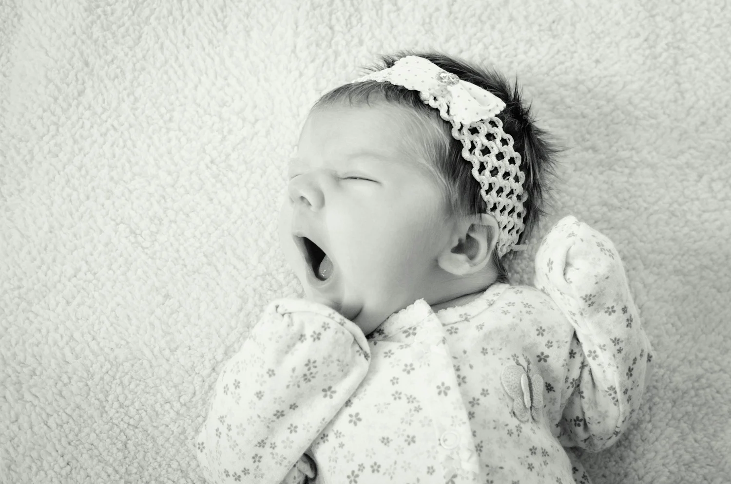 A baby girl yawning while lying on a soft textured blanket, wearing a floral onesie and a lace headband with a bow.
